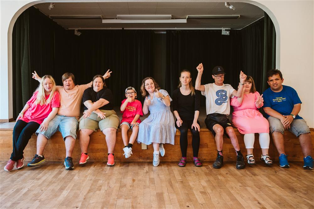 Nine individuals appearing to have different intellectual and motor disabilities seated on a stage in an empty hall.