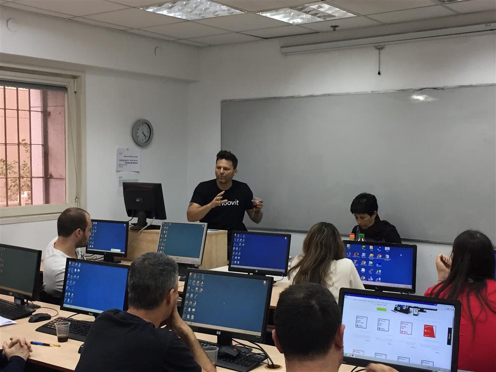 A male and female trainer are at the front of a classroom, which has individual computers on each desk. Five participants sit at the computers, watching the trainers.