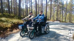 Two persons using their mobility tricycles on a path in the woods.