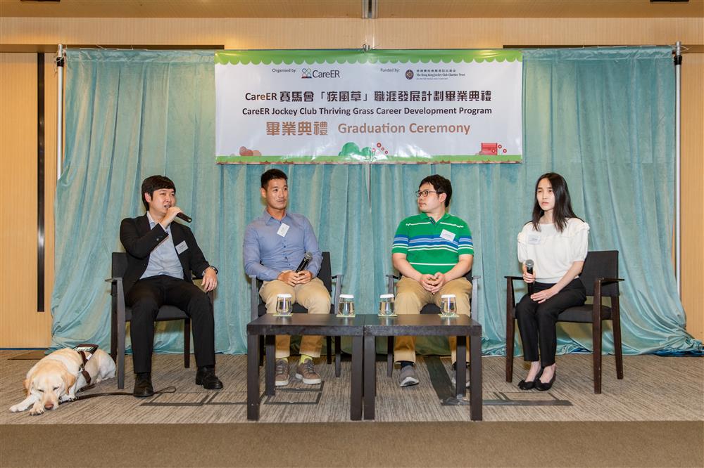 There is a table on a stage with four chairs. Walter Tsui sits on the left with a hand-held microphone and a guide dog beside him. Two men and a woman are in the other chairs.