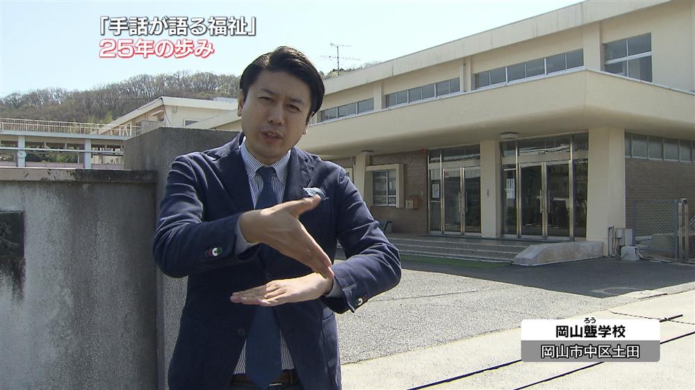 A male tv presenter in a dark blue suit is conveying the news via sign language on television. He is standing outside of a large two story building.