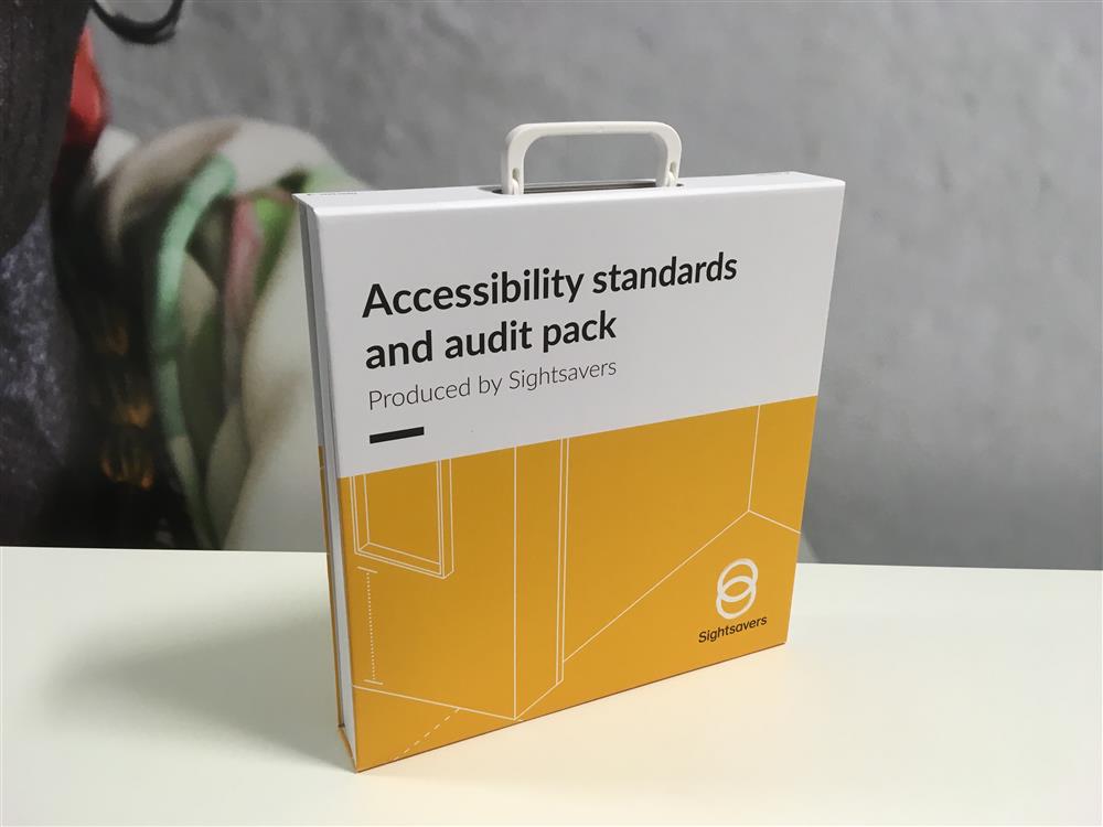 On the edge of a white table there stands a yellow and white information pack with a small white handle. Written on it are the words: "Accessibility standards and audit pack. Produced by Sightsavers."