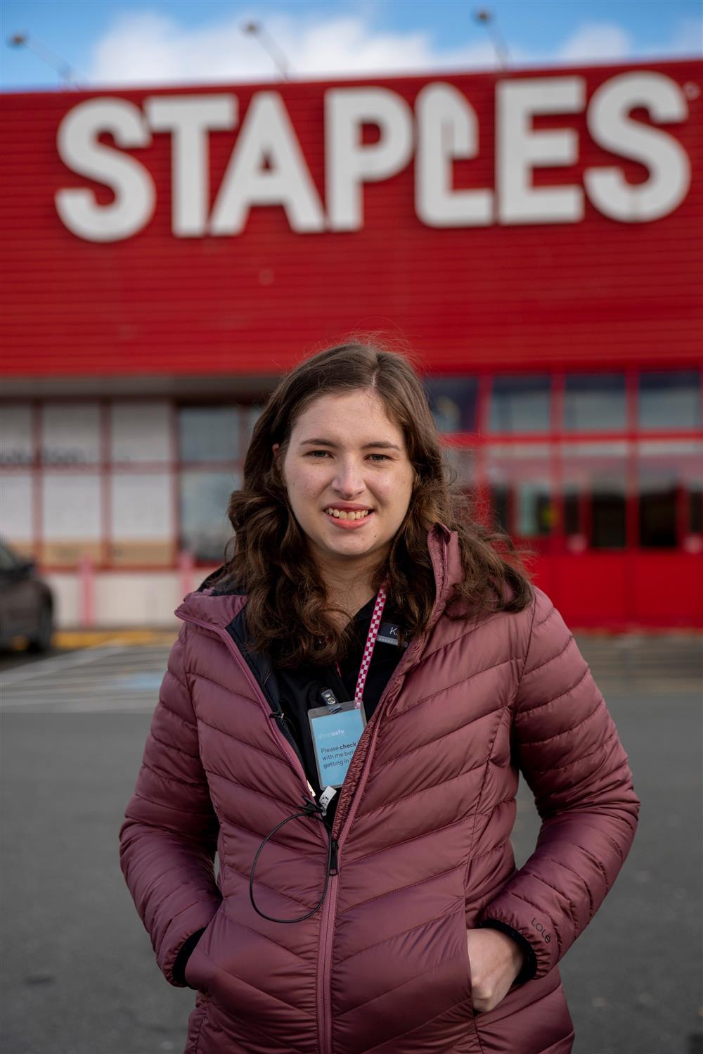 A young woman stands in a car park wearing a maroon jacket and black polo shirt. She is wearing a staff lanyard and looking at the camera and smiling. Behind her is the storefront of Staples. 