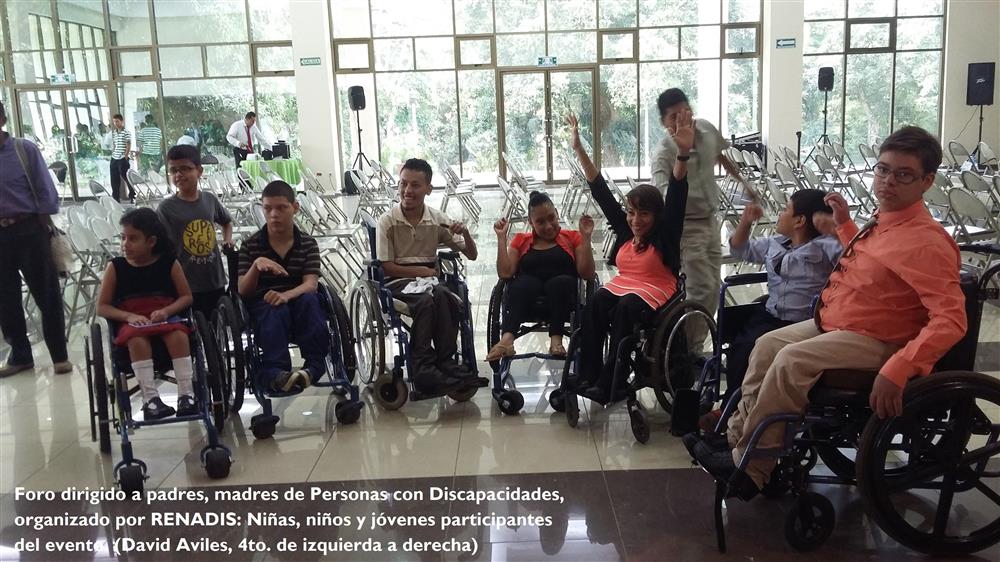 Seven children in wheelchair dressed in formal wear and two teen-aged boys standing behind them inside a big hall with floor to ceiling glass windows at day time.