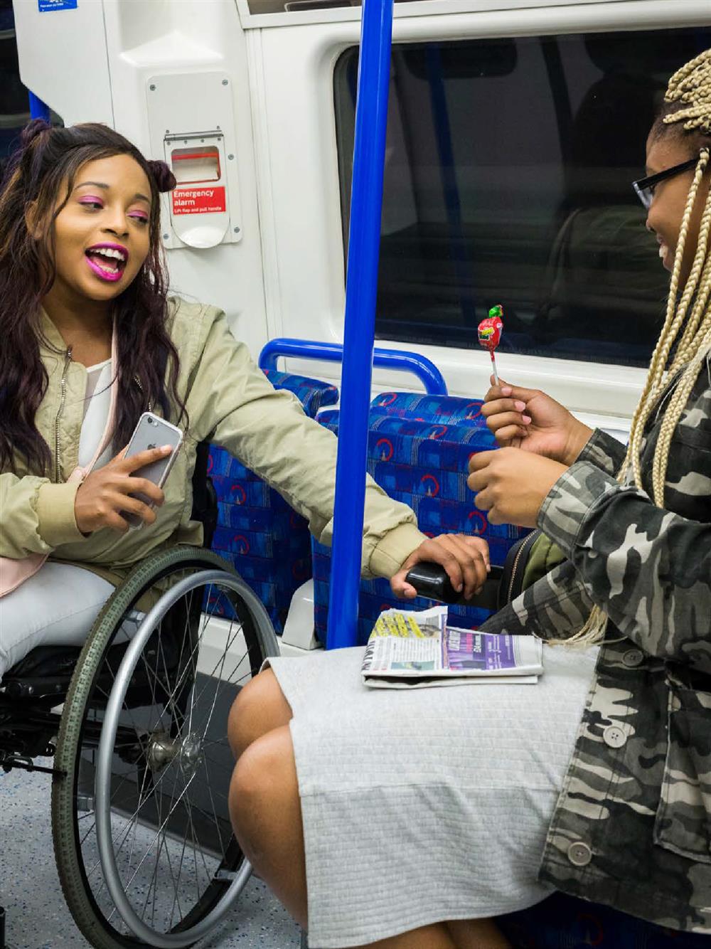 Two women, the one on the left is sitting in a wheelchair, are enjoying a ride on the London tube, conversing and laughing with eachother. The woman on the right is holding a red lollipop. 
