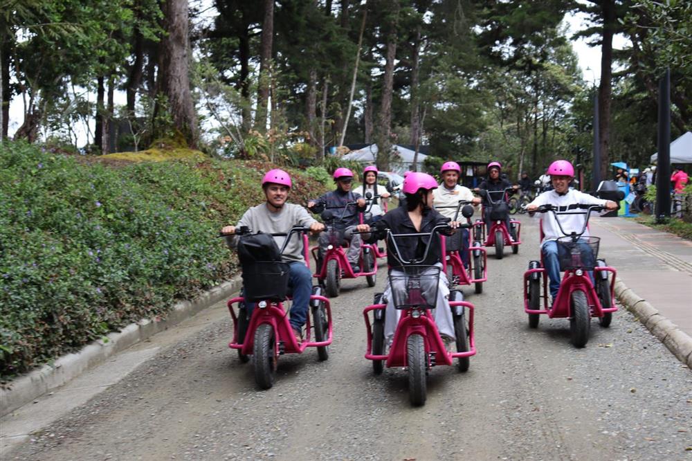 A joyful group of people rides adaptive tricycles through a park, wearing pink helmets. The image radiates freedom, inclusion, and shared recreation, showing how accessible outdoor activities foster community and well-being for everyone.