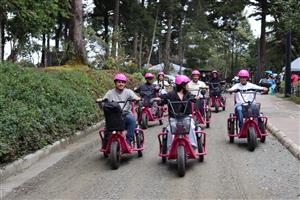A joyful group of people rides adaptive tricycles through a park, wearing pink helmets. The image radiates freedom, inclusion, and shared recreation, showing how accessible outdoor activities foster community and well-being for everyone.
