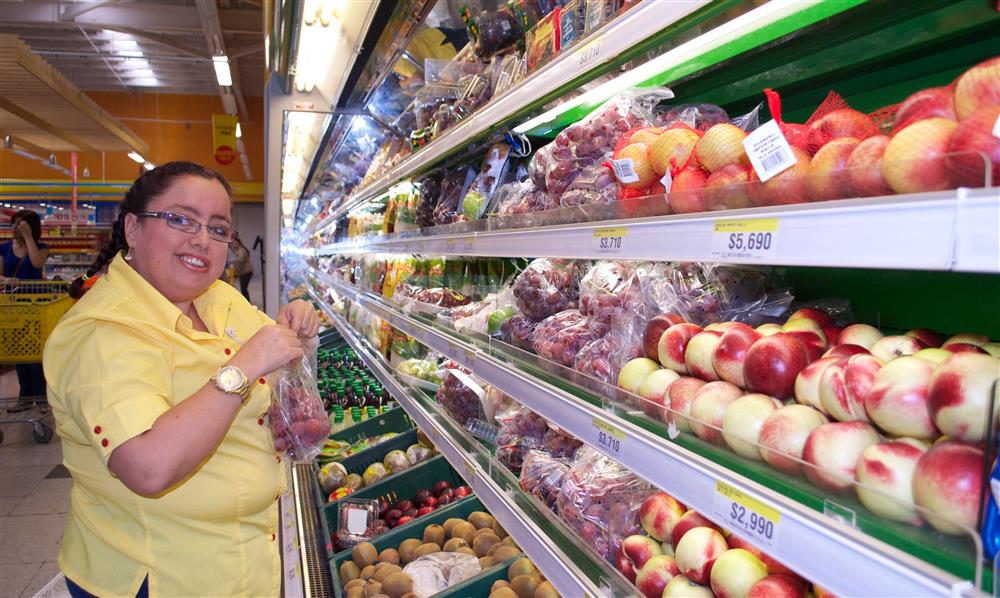 A young woman with dark hair and glasses stands in a supermarket in front of refrigerated shelves of fruit. She is wearing a yellow shirt and holding a bag of red grapes. She is smiling at the camera.