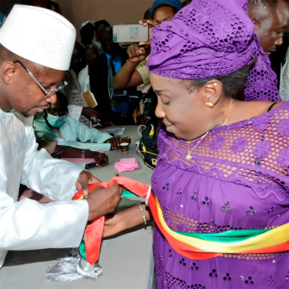 An African man in traditional costume tying a rainbow-colored belt around the waist of a woman also wearing a traditional costume.