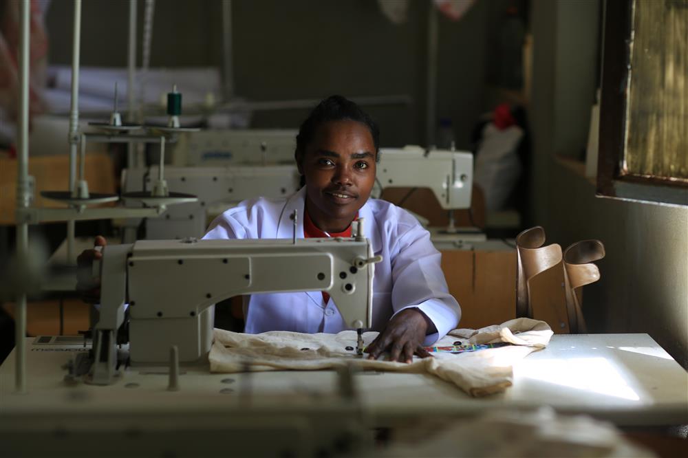 A young woman sits behind a sewing machine, which is placed on a large table. She is feeding material through the machine and looking at the camera and smiling. Crutches lean on the table beside her.