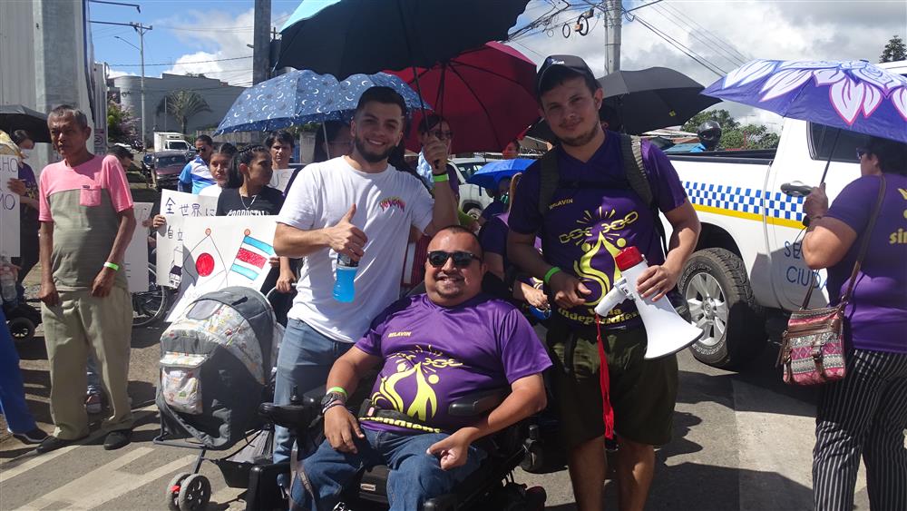 A man seated in wheelchair together with a man holding an umbrella and another man standing behind him in the middle of a street. Other people are walking behind them with placards in what seems to be a mass mobilization.
