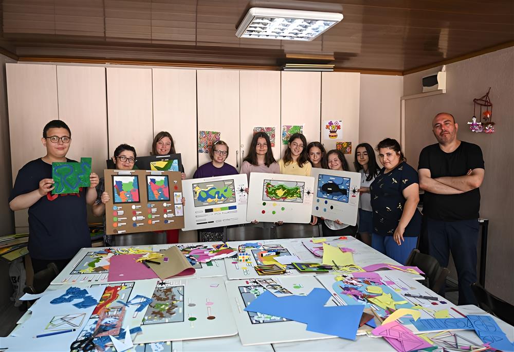 This image shows a group of people, likely a mix of students and instructors, standing proudly in a room filled with colorful artwork. They are displaying their creative work, which includes various abstract designs and shapes. The table in front of them is scattered with paper cutouts and art materials, suggesting a collaborative and educational environment. The diversity in age and the shared focus on art convey a sense of inclusion and community. The setting appears to be an art class or workshop, emphasizing creativity and expression as a unifying activity, promoting values such as teamwork, learning, and the joy of creation.
