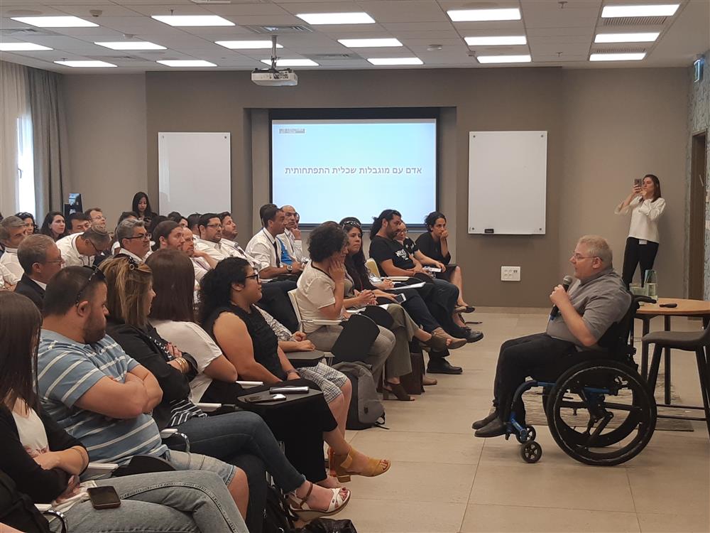 In a bright conferece room with a beamer and a screen in the background, a man in a wheelchair holds a microphone and speaks to a large attentive audience, sitting on chairs. 