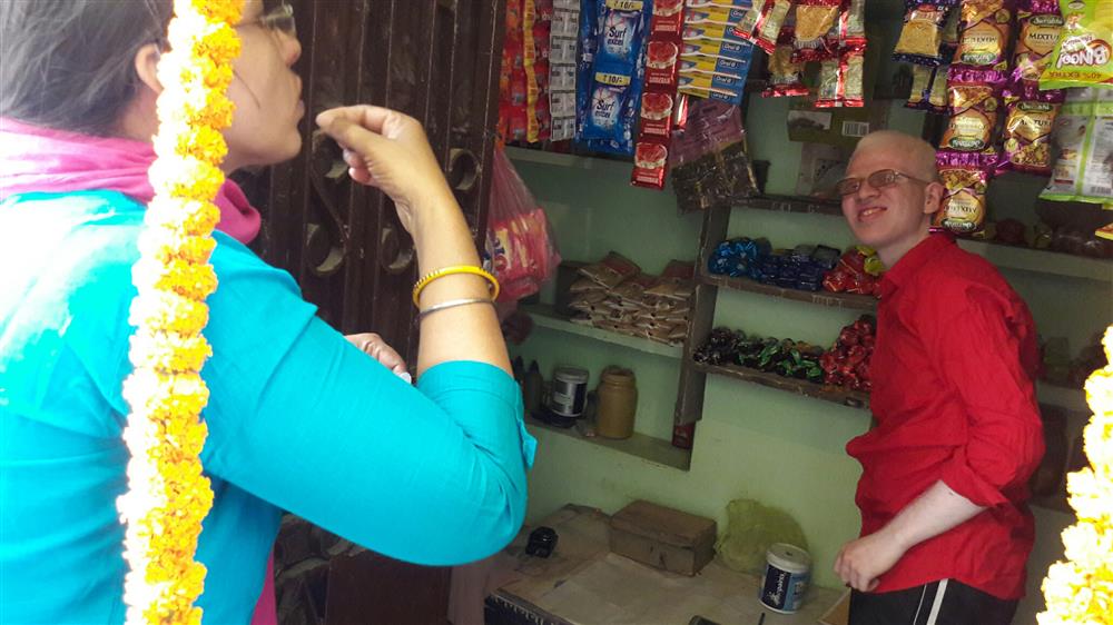 A man stands in a small shop. Behind him are shelves with snacks and household products. He is smiling at a woman standing in the shop doorway who is communicating with him in sign language.