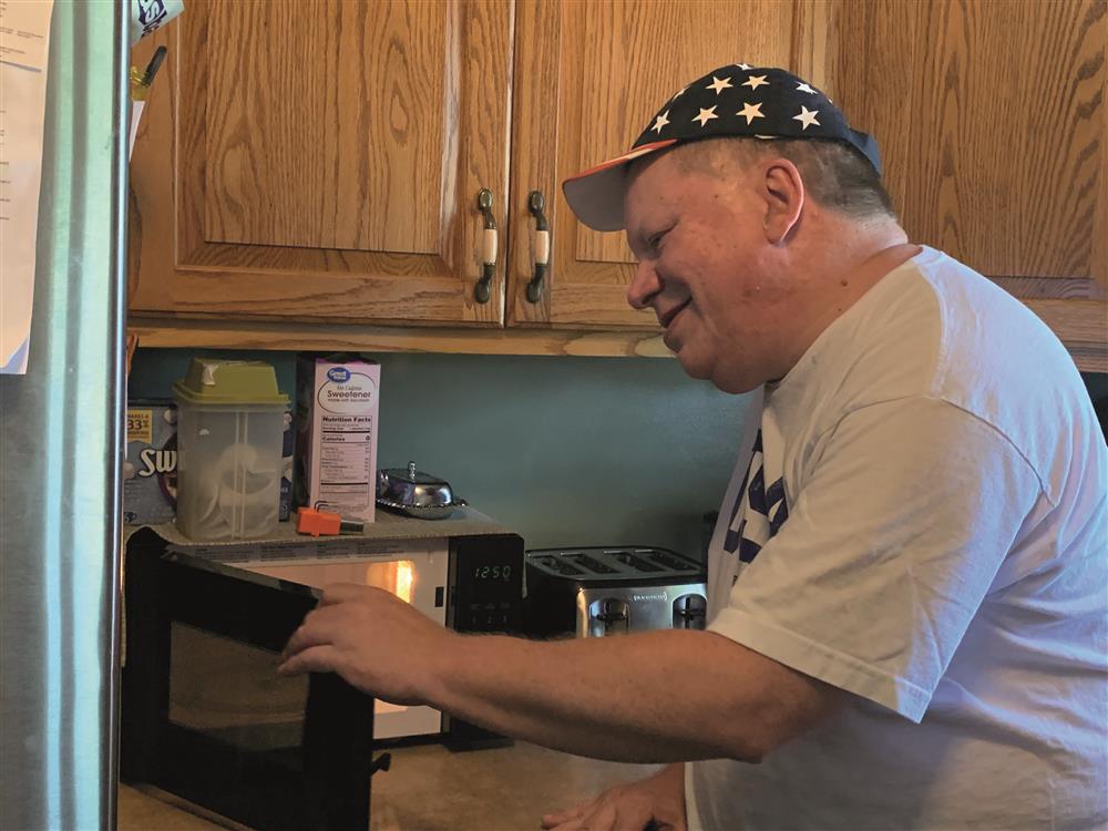 A middle-aged man who appears to have mental disability dressed in shirt and baseball cap is taking something from the microwave in the kitchen.