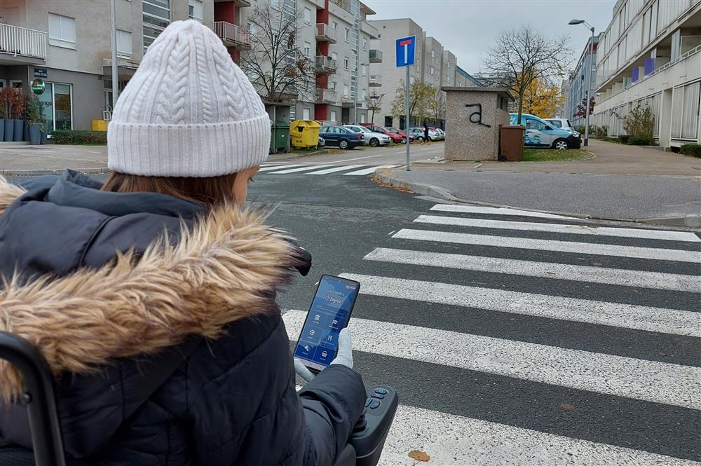 A young woman sitting in her electric wheelchair looks at the "Accessbile Zagreb service" app on her smartphone. She is waiting in front of a Zebra crossing in a residential area.  