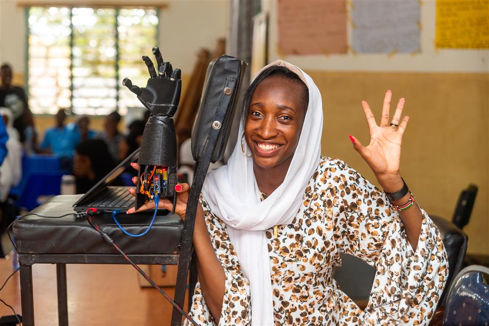 A young African woman smiles proudly beside a robotic prosthetic hand connected to a laptop. Her confident pose and enthusiasm celebrate innovation, education, and empowerment—showing how technology can enhance accessibility and equality in science and engineering.