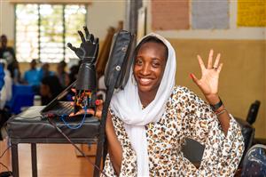 A young African woman smiles proudly beside a robotic prosthetic hand connected to a laptop. Her confident pose and enthusiasm celebrate innovation, education, and empowerment—showing how technology can enhance accessibility and equality in science and engineering.