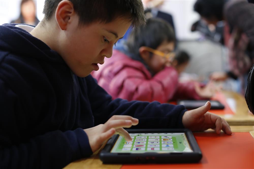A young boy with down syndrom is very engaged in playing on a tablet while at the same time expanding his vocabulary. In the background other children do the same. 