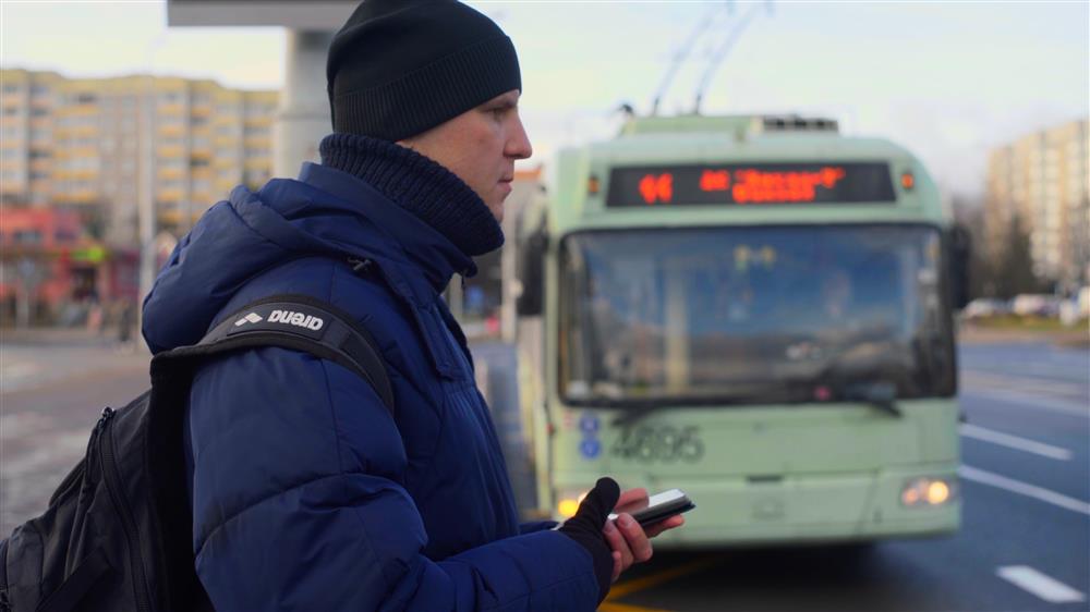 A man dressed warmly waits for a bus on a cold city morning, holding his phone and backpack. The photo highlights the importance of accessible and reliable public transportation, essential for inclusion and equal mobility in urban environments.