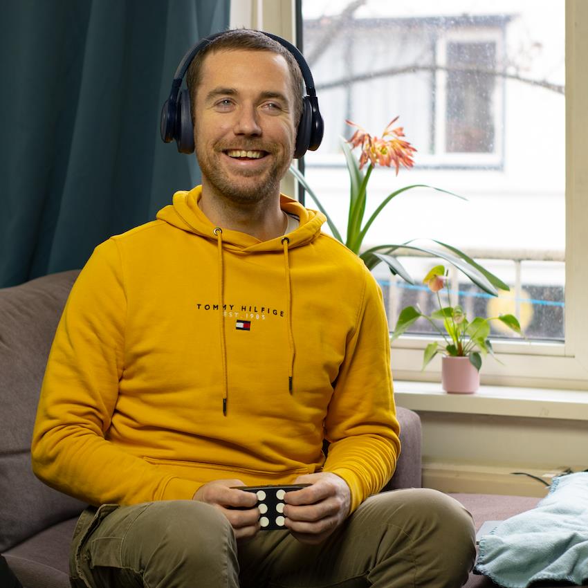 In the image, we see a person sitting comfortably on a couch, wearing a vibrant yellow hoodie and headphones, with a joyful smile on their face. They are holding what appears to be a video game controller. The setting is a cozy room with a window through which daylight is coming in, and there are plants on the windowsill, adding a touch of nature and tranquility to the scene. The atmosphere is relaxed and the individual seems to be enjoying their leisure time, which may suggest a balance between work and personal life, an important aspect of well-being and equality.