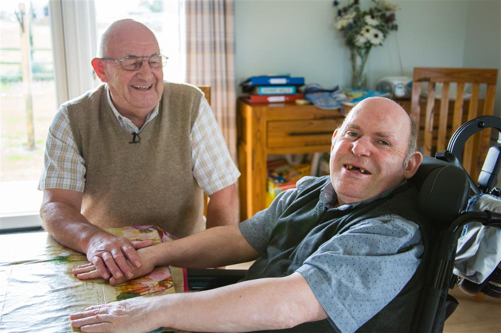 Two men, one a wheelchair user, sitting in a living room and smiling