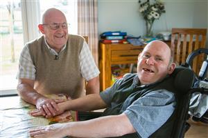 Two men, one a wheelchair user, sitting in a living room and smiling