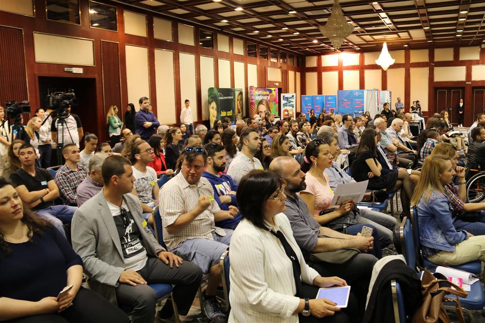 Many women and men are sitting in rows, looking at a podium; in the background are promotional stands.