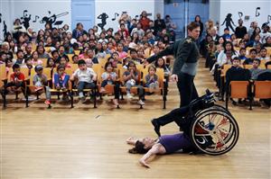 This image shows a diverse group of schoolchildren watching a performance in a school auditorium. In the foreground, a person in a wheelchair appears to be engaging in an expressive dance or performance art, while another performer lies on the floor, reaching out. The audience looks captivated and surprised by the performance. The scene promotes inclusivity and the idea that art and expression are accessible to all, regardless of physical abilities. The performers demonstrate courage and creativity, which serves as an inspiration to the audience, highlighting themes of equality and the celebration of diverse abilities.
