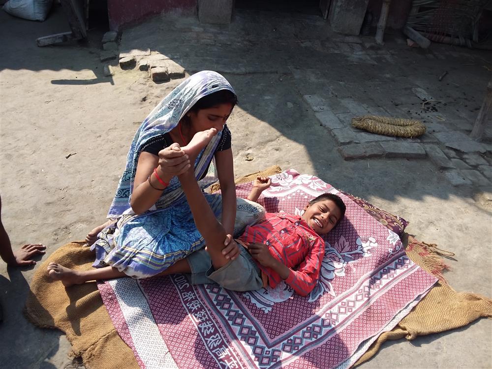 An Indian woman wearing a blue Sari is stretching the legs of a young boy with a physical disability who is laying on two layers of colored cloth and smiling into the camera. 