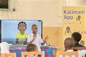 A student presents confidently in front of classmates, demonstrating sign language beside a display introducing the “Kalimani App.” This scene celebrates African innovation and accessibility in education, promoting communication and inclusion for Deaf learners.