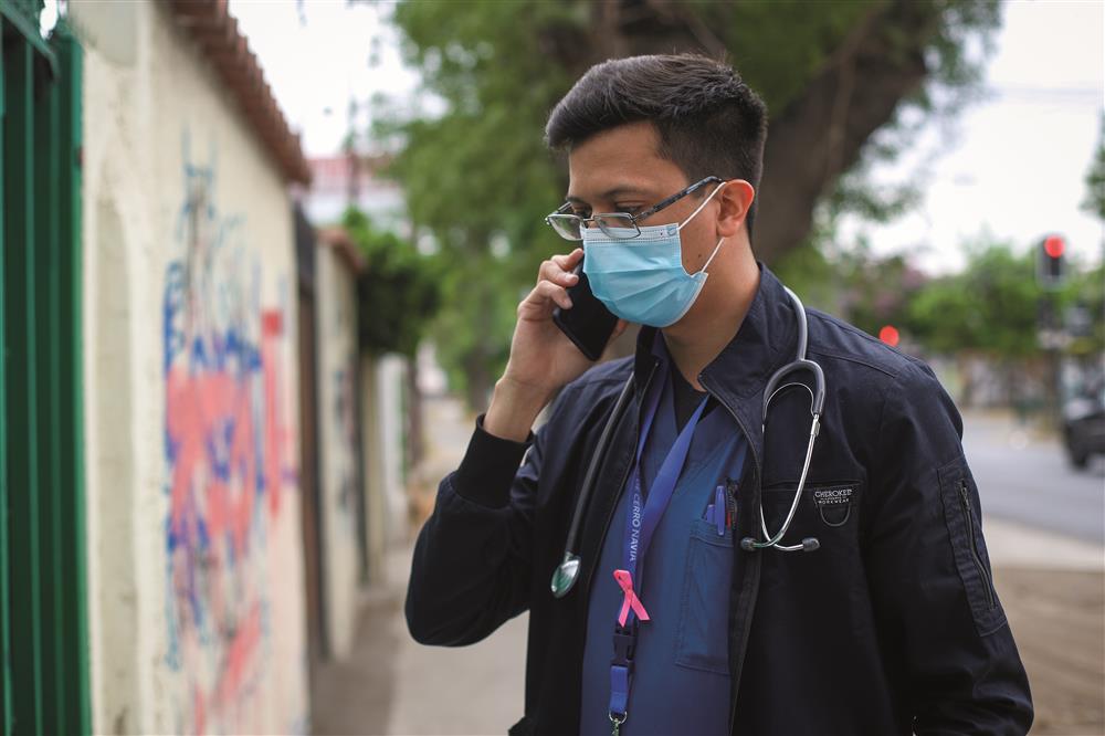 A man wearing a surgical mask, a black jacket, a blue lanyard with pink ribbon and a stethoscope around his neck is holding a phone on his right ear and standing in a sidewalk facing a house.