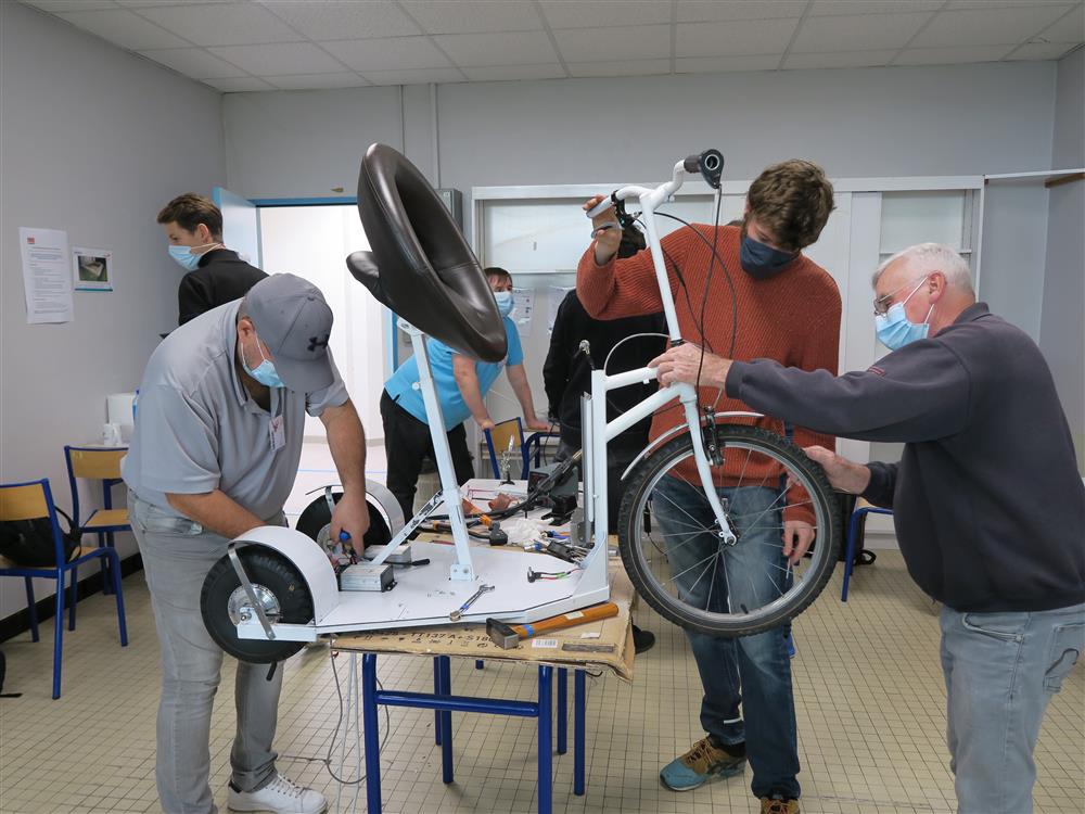 Three men with masks work on a three-wheel bicycle. Two work at the front and one fixes something in the back. They are in a working environment 