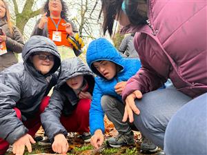 Children and young people in warm jackets explore nature closely on a rainy day, guided by caring adults. Their curiosity and teamwork reflect the importance of accessibility in outdoor education—where every child can participate, learn, and connect with the world.