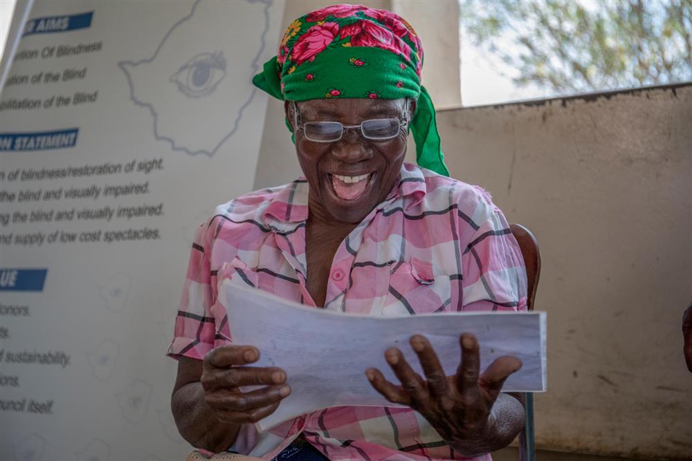 An elderly African woman beams with joy as she reads a document, wearing new eyeglasses and a bright green headscarf. Her laughter conveys empowerment and dignity, symbolizing how access to vision care restores not just sight, but independence and confidence.