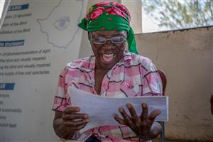 An elderly African woman beams with joy as she reads a document, wearing new eyeglasses and a bright green headscarf. Her laughter conveys empowerment and dignity, symbolizing how access to vision care restores not just sight, but independence and confidence.