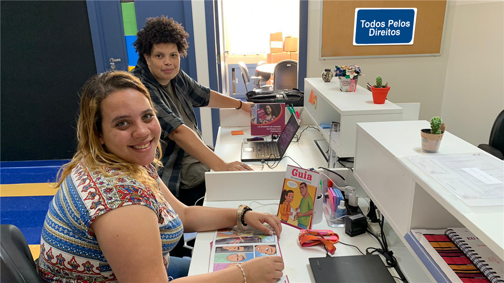 Two Brazilian ladies seated on a working desk beside each other in an office room. A corkboard has a signage "Todos Pelos Diretos" on the wall perpendicular to the ladies.