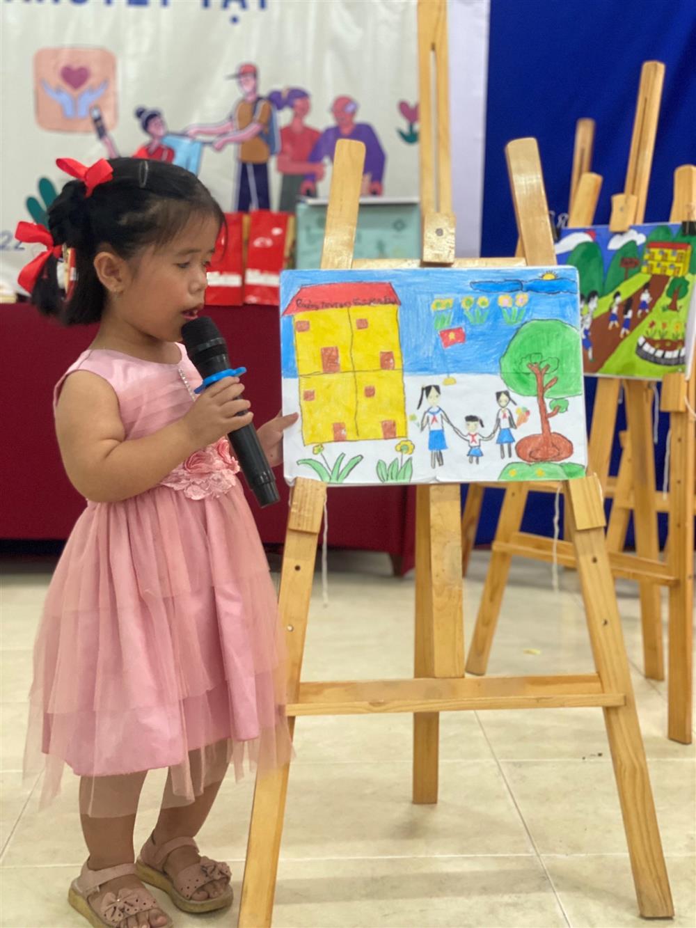 The photo shows a young girl in a pink dress, standing in front of an easel and speaking into a microphone. The easel displays a colorful drawing that appears to be created by a child, featuring a yellow building, greenery, and figures that could represent people. In the background, there are additional easels with artwork, suggesting this may be an art exhibition or school event. The setting promotes creativity and expression, and the girl's participation indicates a supportive environment fostering confidence and public speaking at a young age.