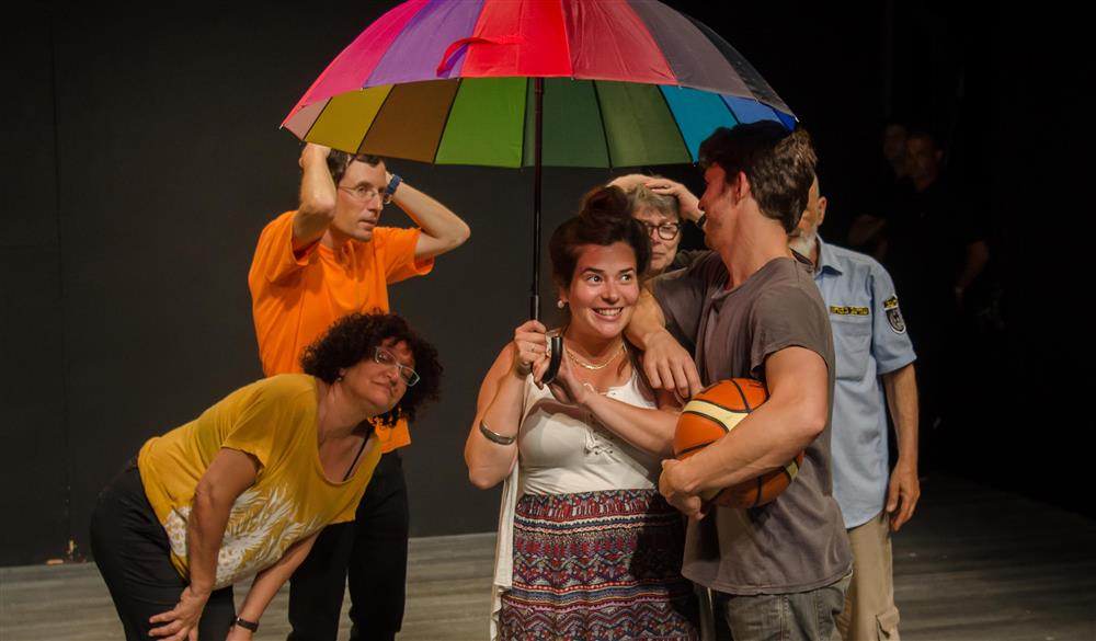 A theatre group of 6 people stands under a rainbow colored umbrella.