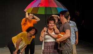 A theatre group of 6 people stands under a rainbow colored umbrella.