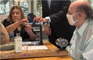 An elderly woman and man, the latter wearing a face mask, sit at a table while a younger person helps them with a tactile art activity. The scene reflects intergenerational respect and inclusive creativity, supporting active participation regardless of age or ability.