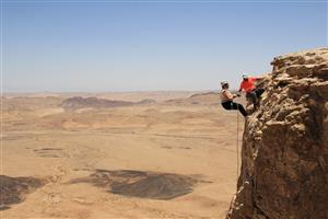 On the right side of the picture there is a man helping a woman lowering herself down on a rope form a large brown rock. In the background there lies a mountainous and desertlike landscape stretching until the horizon. 