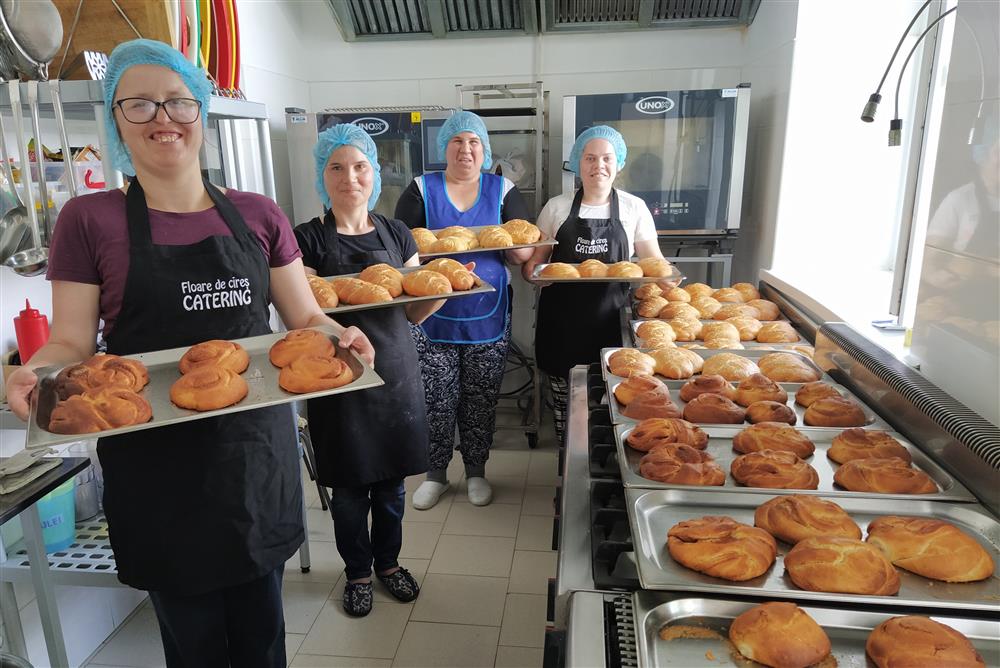 Four women in work uniforms hold trays with bakery products in front of them. There are ovens in the background and more trays with bakery products on the side