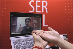 A person's right hand placed on top of the left hand with red bracelet in front of a laptop. On the laptop screen is a bearded man seated with background "SERVEL" printed his thumb pointing up on the screen.