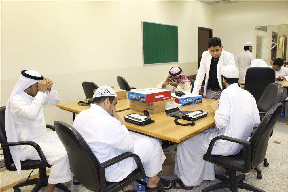 A group of 6 young male students dressed in white thobes sit in a classroom and test out braille notetakers positioned on a brown table in front of them while a teacher is standing beside. 