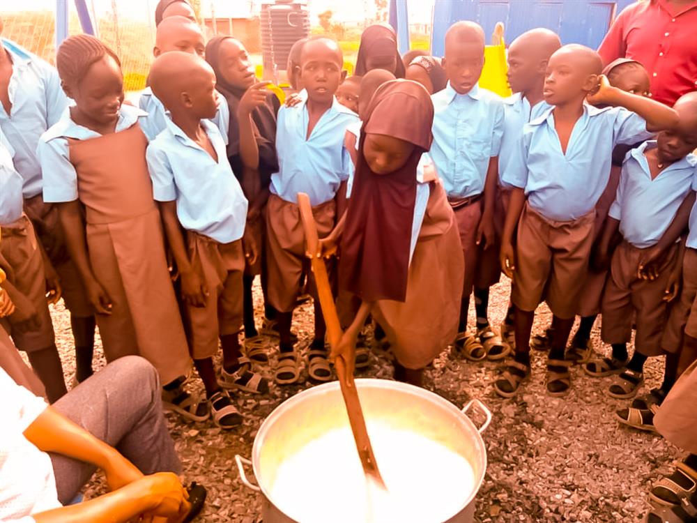 The photo depicts a group of African children in school uniforms, standing around a large cooking pot outdoors. One child, who is wearing a hijab, is stirring the pot with a long wooden spoon, suggesting a communal activity or shared meal. The children appear engaged and curious about the process. The image conveys a sense of community, cooperation, and inclusivity, as children from different backgrounds come together in a shared experience. It also subtly touches on the importance of providing assistance and ensuring that all members of a community, including children, have access to basic needs such as food.