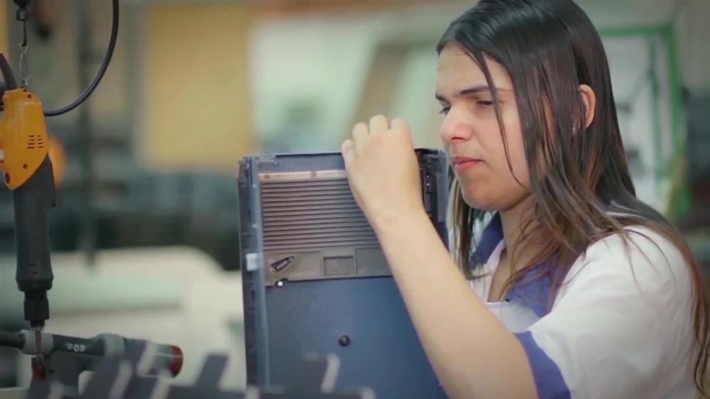 A young woman with dark hair and wearing white overalls is at a workstation in a workshop. She is handling a metal casing and a drill is attached to the workspace in front of her.