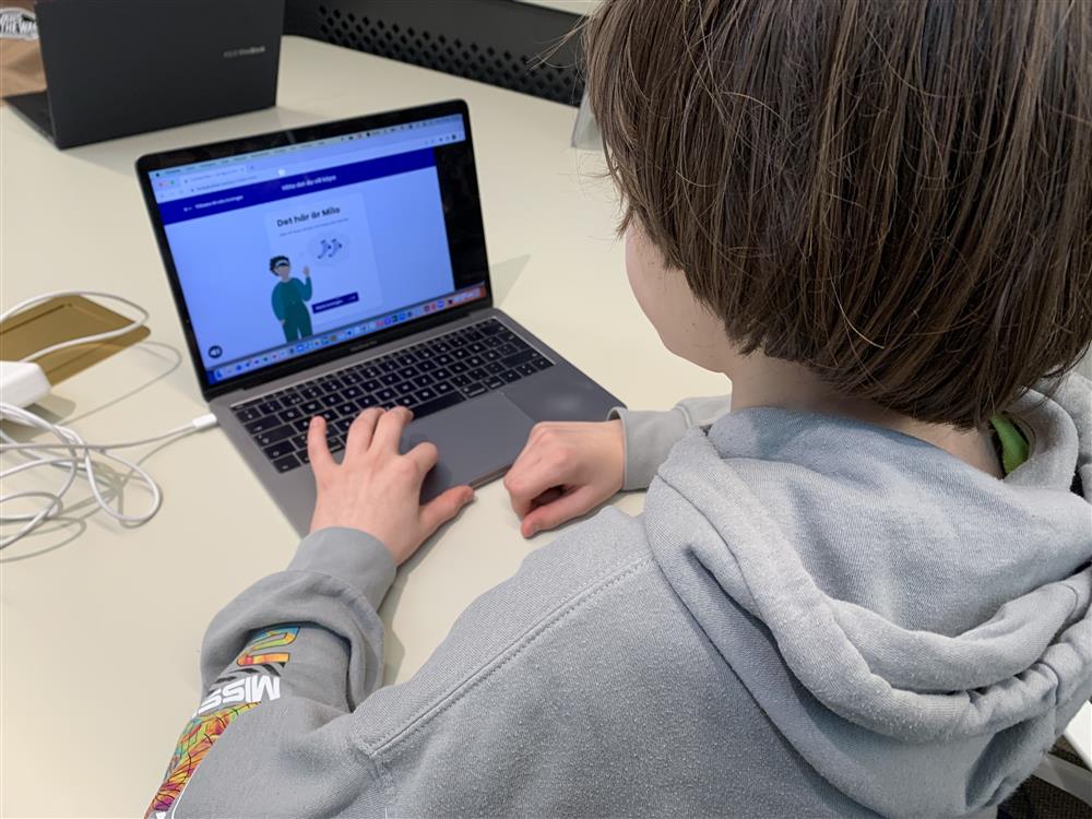 A boy in hoodie looking at his computer. In his screen is an animation of a boy and a pair of socks with a banner "Det här är Milo".