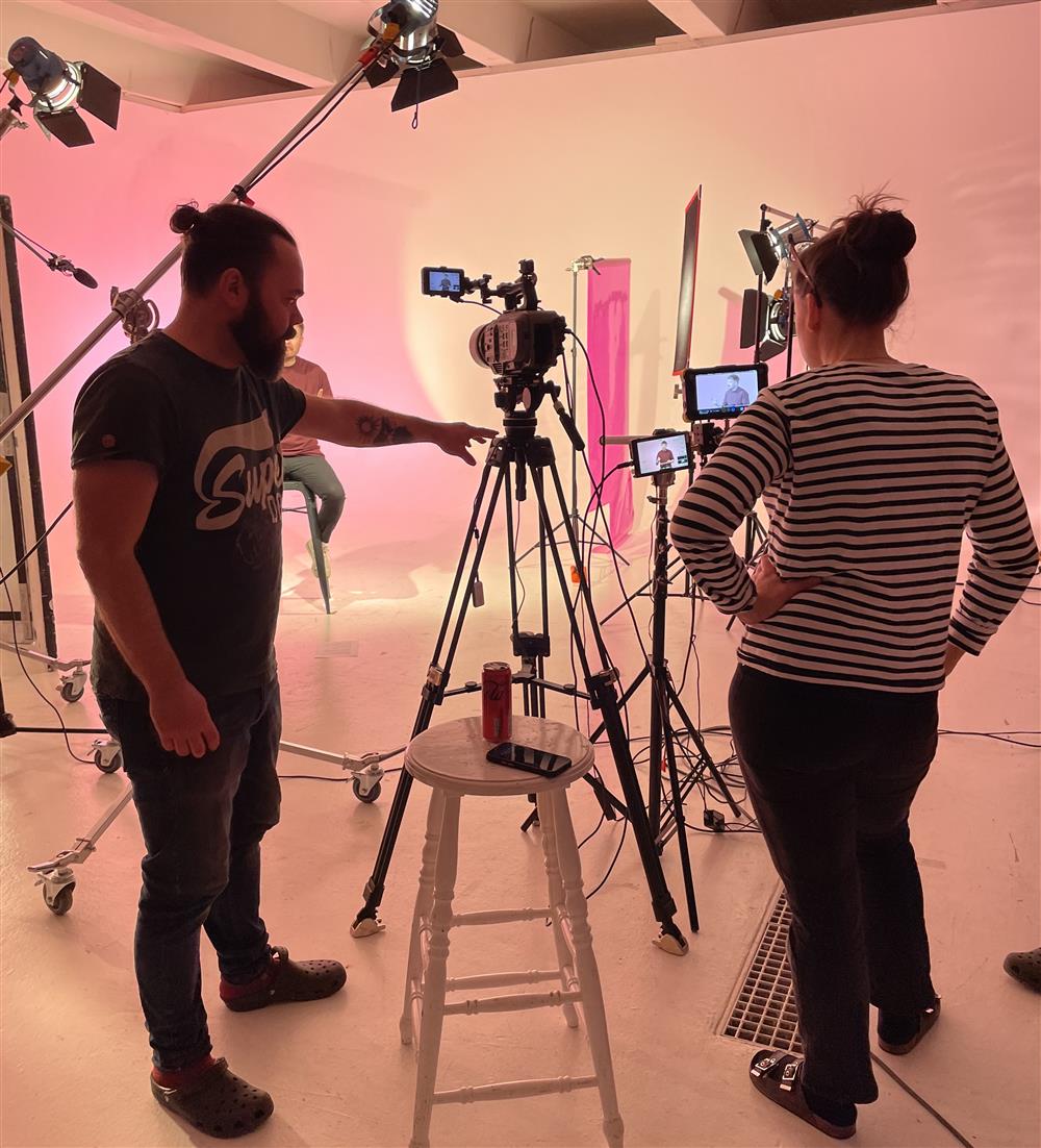 In a warmly lit studio, two people work behind cameras, adjusting lighting and angles during a film shoot. The teamwork and creativity reflect collaboration in inclusive storytelling—where diverse voices and perspectives are represented on screen.