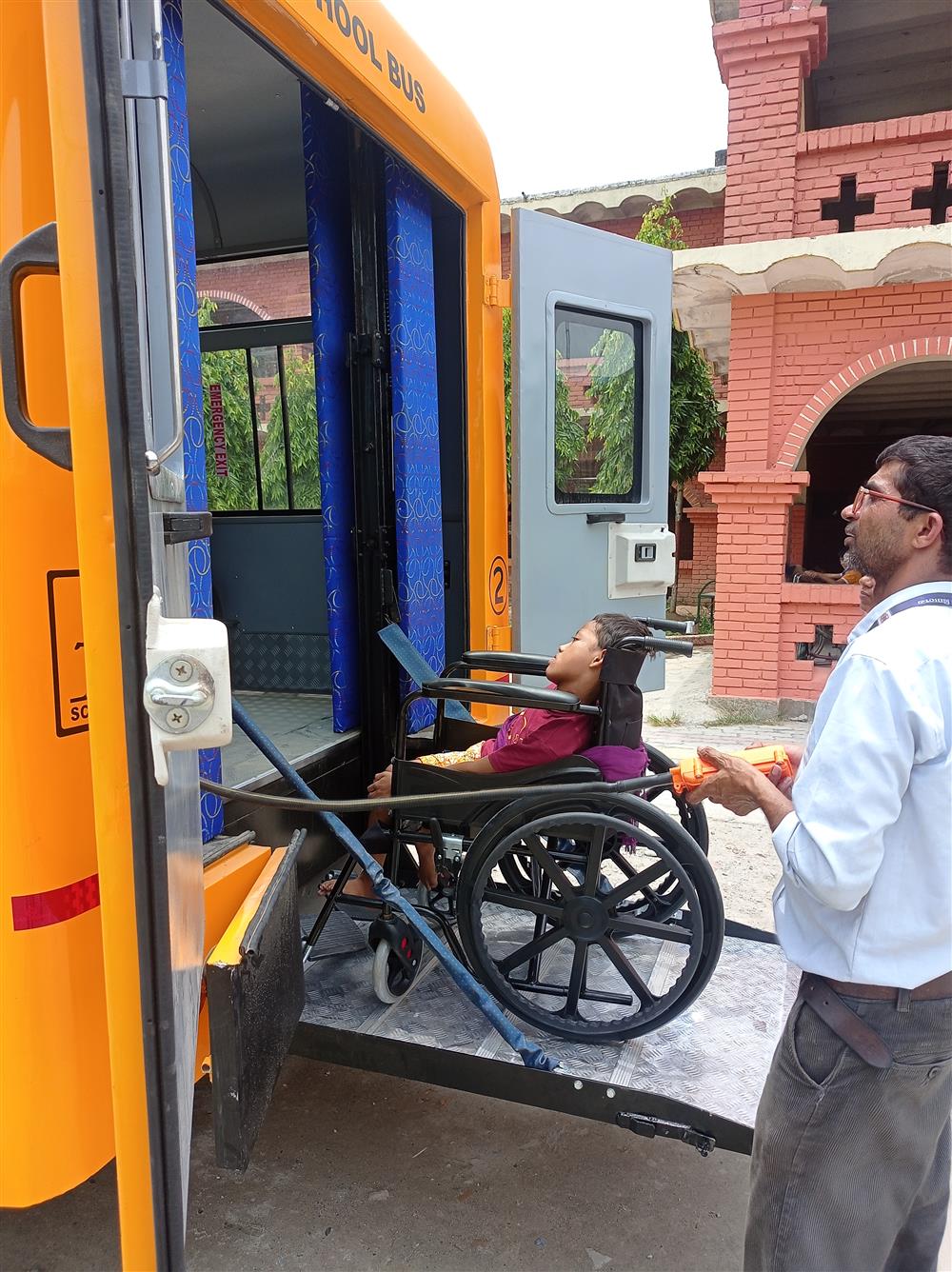 The photo shows a person in a wheelchair being assisted onto a school bus equipped with a ramp, highlighting accessibility for individuals with disabilities. A man is seen operating the ramp, ensuring the safe boarding of the wheelchair user. The bus is brightly colored, and the setting appears to be outside a building with red brickwork. This image reflects a commitment to inclusivity and the importance of providing equal opportunities for mobility and education to all members of society.